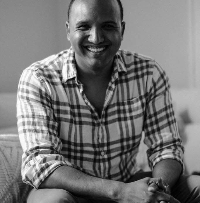 Black and white portrait of a smiling man wearing a plaid shirt with hands clasped.