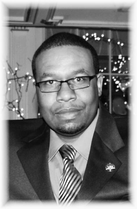 Black and white portrait of a man with glasses, goatee, suit, and striped tie.