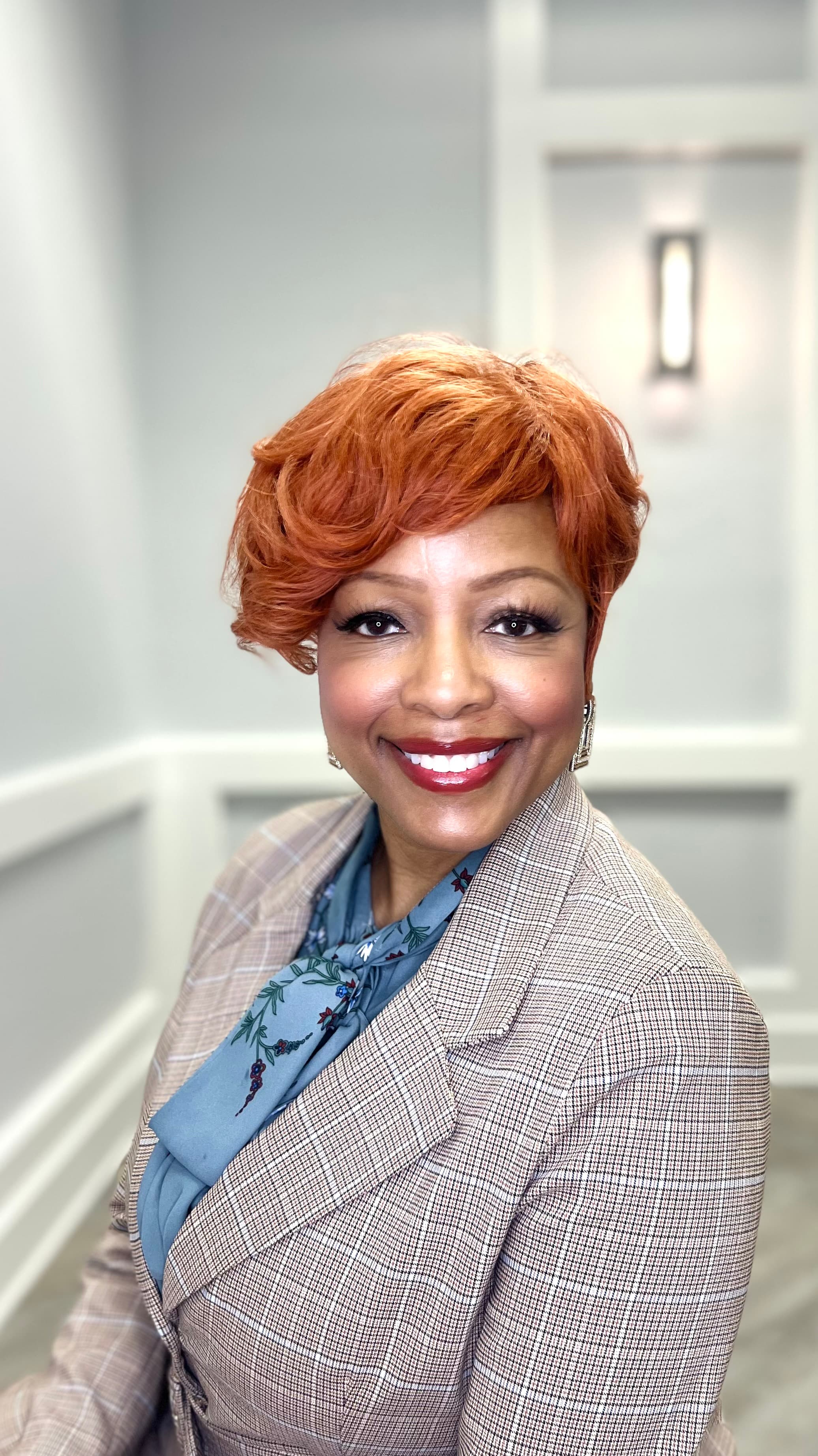 Smiling woman with short copper hair wearing a tan blazer and blue floral blouse.
